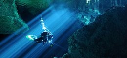 Light beams shine on a static diver surronded by rock formations in a cenote.